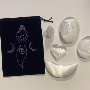 Puede incluir: Un conjunto de seis cristales de selenita blancos, que incluyen un corazón, una luna creciente, un cristal redondo con un diseño de árbol, un cristal redondo con un diseño de triple luna, una punta y una bolsa de terciopelo negro con un diseño bordado en morado de una mujer sosteniendo una esfera.