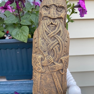 Puede incluir: Una estatua tallada de color marr&oacute;n dorado de un hombre barbudo con una espada. La estatua est&aacute; de pie sobre una superficie blanca con una superficie azul detr&aacute;s. Hay flores moradas en el fondo.
