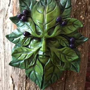 May include: A green leaf mask with a face carved into it, featuring a detailed design of leaves and berries. The mask is mounted on a wooden surface, showcasing a rustic and natural aesthetic. The face has a serene expression.
