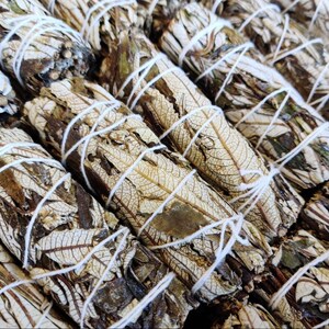 May include: Close-up of a bundle of white sage smudge sticks tied with white twine. The sticks are made of dried sage leaves and are arranged in a pile.