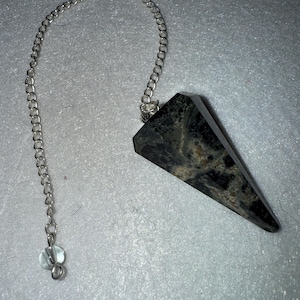 May include: A black and brown crystal pendulum with a silver chain and a small clear bead at the end. The pendulum is a dark, faceted stone with a pointed tip.
