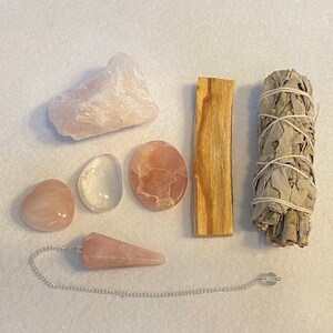 May include: A set of pink rose quartz crystals, a clear quartz crystal, a palo santo stick, and a bundle of white sage. The set also includes a pink rose quartz pendulum with a silver chain.