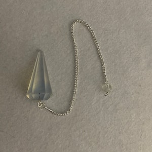 May include: A clear crystal pendulum with a silver chain. The pendulum is shaped like a teardrop and has a pointed tip.