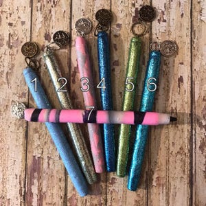 May include: Seven glitter pens in various colors, including blue, silver, pink, and green, are arranged on a distressed wooden surface. Each pen has a decorative charm attached to the top. The pens are numbered 1 through 7.