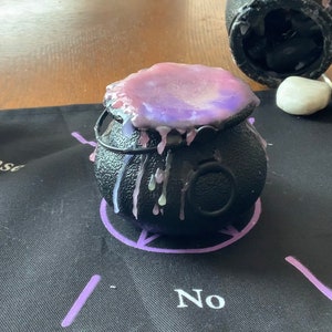 Puede incluir: Un caldero negro con una vela de cera morada y blanca en su interior. El caldero está sentado sobre una superficie de tela negra con un círculo morado y la palabra "No" escrita en blanco.