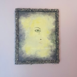 May include: A framed painting of a stylized face with a single teardrop falling from the eye. The background is a muted yellow and gray with a textured look. The frame is made of small, gray stones.