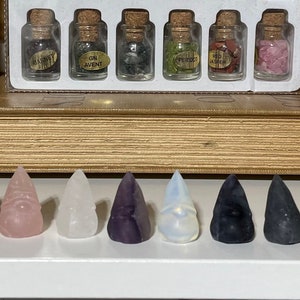 May include: A row of 10 small, pointed crystal shapes in various colors, including pink, blue, purple, white, black, and grey. The crystals are arranged on a white shelf.