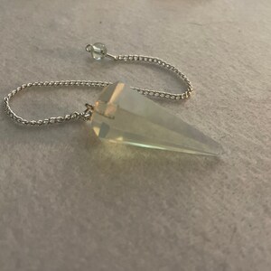 May include: A clear quartz crystal pendulum with a silver chain. The pendulum is a pointed cone shape.