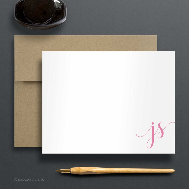 May include: A white card with a pink cursive monogram "js" in the bottom right corner. The card is on a brown envelope and a wooden pen is in front of it.