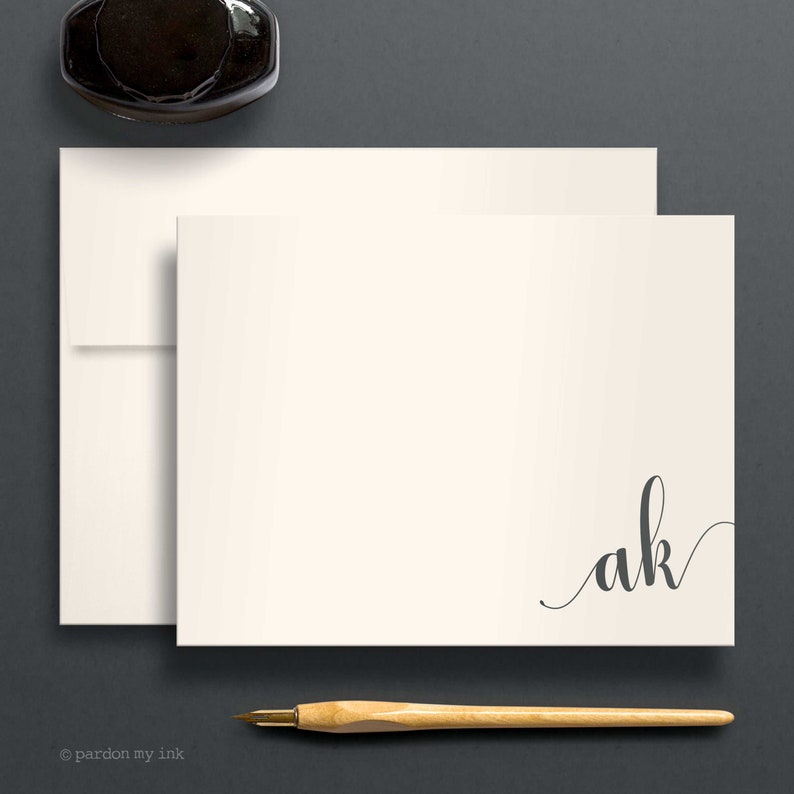 May include: A white card with a grey cursive monogram "ak" in the bottom right corner. The card is on a grey surface with a black inkwell and a wooden pen.