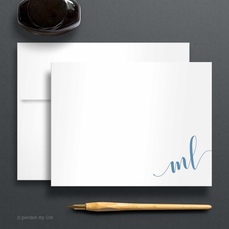 May include: Two blank white stationery cards with a blue cursive monogram "ml" in the bottom right corner of the card. A wooden pen is lying on a grey surface below the cards.