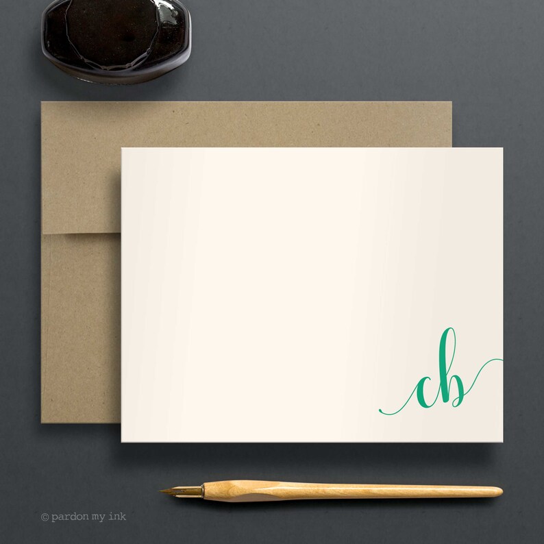 May include: A white card with a brown envelope and a green calligraphy monogram "cb" in the bottom right corner. A wooden pen is lying on a grey surface.