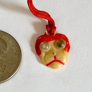 May include: A small, vintage pendant shaped like a face with red hair and a red string. The face is off-white with two round, light-colored eyes. A U.S. quarter is next to the pendant for size comparison.