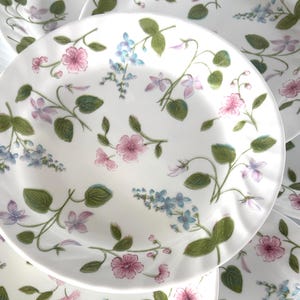 May include: A set of white ceramic plates with a floral design. The plates are decorated with pink, blue, and purple flowers, along with green leaves and stems. The plates have a slightly scalloped edge.