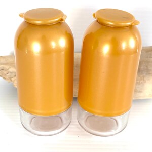 May include: Two cylindrical containers with metallic gold-colored upper sections and clear glass bases. Each container features a matching gold-colored circular lid on top. The containers are positioned side-by-side, displaying a smooth finish.