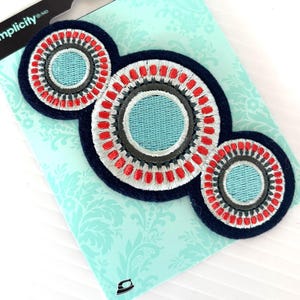 May include: Embroidered applique patches in a circular design with red, gray, and light blue details. The patches are on a light blue card with a floral pattern. The brand name "Simplicity" is visible.