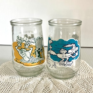 May include: Two glass jars with cartoon characters from the Looney Tunes. The jar on the left features Wile E. Coyote falling into a hole with the Road Runner running away. The jar on the right features Wile E. Coyote on roller skates with a rocket strapped to his back. The text "ACME" is on the rocket.