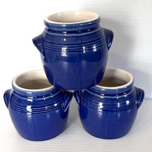 May include: Three cobalt blue ceramic pots with a cream-colored interior. Each pot has two small handles and a ridged design around the middle. The pots are stacked in a triangular arrangement against a white background.