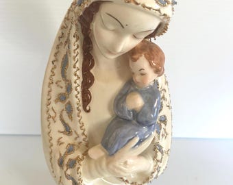 May include: A ceramic figurine depicting a mother holding a child. The mother wears a cream-colored veil with gold and blue accents. The child is dressed in blue. The base is cream-colored with a textured design.
