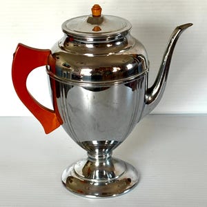 May include: A polished chrome teapot featuring a vibrant orange handle and a small orange knob on the lid. The teapot has a rounded body with vertical fluted detailing, a long curved spout, and a circular pedestal base for stability.