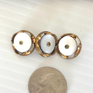 May include: Vintage brooch featuring three white, circular, mother-of-pearl cabochons set in ornate, gold-toned metal frames. The brooch is approximately 2 inches long and is next to a quarter for size comparison.