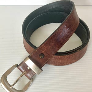 May include: A brown leather belt with a silver buckle. The belt is folded in half and the buckle is facing the camera.