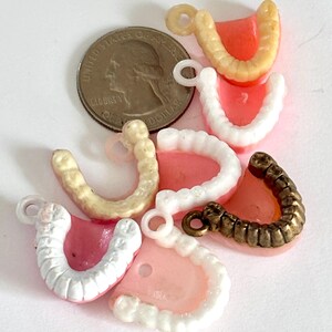 May include: A collection of miniature dentures in various colors, including pink, white, and beige, with a quarter for size comparison. Some dentures have small loops for attaching to jewelry or other crafts.