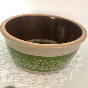 May include: A small, round, brown and white ceramic bowl with a green band and speckled design. The bowl is sitting on a white doily.