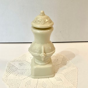 May include: A white ceramic bottle with a floral design on the lid and a square base. The bottle is sitting on a white crocheted doily.