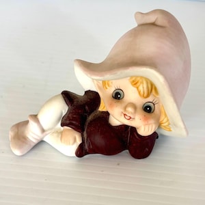 May include: A ceramic figurine of a whimsical elf with a large, light pink hat. The elf has blonde hair, rosy cheeks, and is dressed in a burgundy top and white pants. The elf is lying down, resting its chin on its hand.