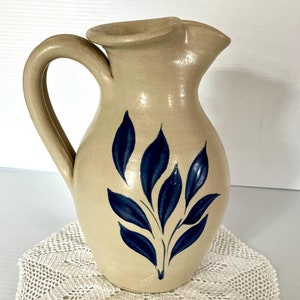 May include: A beige ceramic pitcher with a dark blue leaf design. The pitcher has a handle and a spout. It sits on a cream-colored, crocheted doily. A decorative kitchenware item.