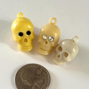 May include: Three small skull charms with loops at the top for hanging. One is bright yellow, one is pale cream with clear rhinestone eyes, and one is off-white. A US quarter coin is placed next to them to show the scale of the charms.