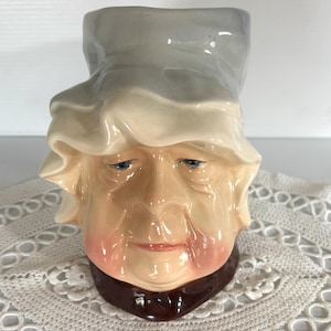 May include: A ceramic mug shaped like a woman's head with a white bonnet. The mug has a brown collar and a light blue bonnet. The woman's face is painted with realistic features, including wrinkles and rosy cheeks.