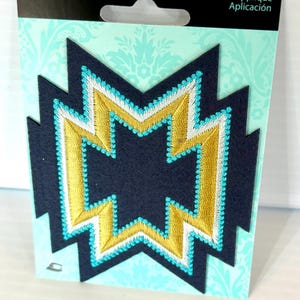 May include: A navy blue appliqué patch with a gold and white geometric design, outlined with turquoise stitching. The patch is on a light blue background with the text "Simplicity" and "Appliqué Application".
