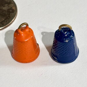 May include: Two small, bell-shaped thimbles, one orange and one blue, with gold-colored loops at the top. The thimbles are next to a quarter coin for size comparison. The thimbles have a textured surface.