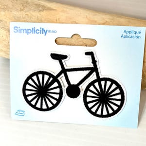 May include: Black bicycle applique on a white background. The applique is on a light blue card with the brand name "Simplicity" and the word "Appliqué" printed on it. The applique is iron-on.