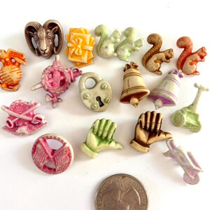 May include: A collection of small, molded decorative buttons in various shapes including a ram head, squirrels, bells, hands, a padlock, and a peace sign, arranged near a US quarter coin for scale, featuring diverse colors like pink, green, and brown.