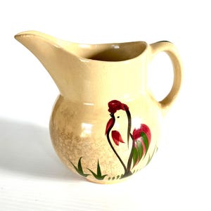 May include: A cream-colored ceramic pitcher featuring a hand-painted rooster design on the front. The rooster is depicted in red, green, and black colors, standing amidst small green grass blades. The pitcher has a curved handle and a wide spout.