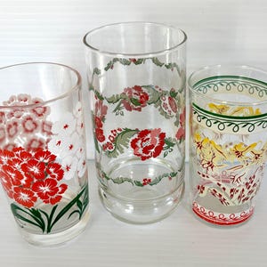 May include: Three vintage clear glass tumblers with floral and decorative designs. One tumbler features red and white flowers, another has a floral garland, and the third has a decorative pattern with red and yellow accents.