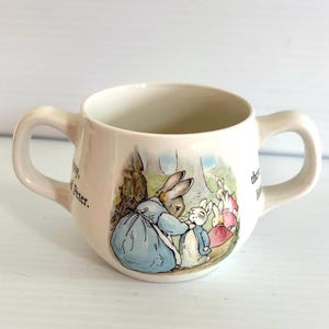 May include: Cream-colored ceramic baby cup with two handles. The cup features a colorful illustration of rabbits in a forest scene. The cup has text on the side and is designed for small children.