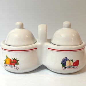 May include: A white ceramic condiment holder with two lidded compartments and a handle. Each compartment features a red stripe and a fruit graphic with the "Smucker's" logo. One side shows an orange and raspberries, the other grapes, an apple, and a pear.