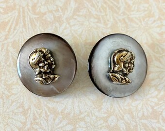 May include: A pair of vintage button earrings with a silver and gold design of a man's head in profile. The buttons are round and have a white and gray mottled appearance.