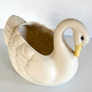 May include: A ceramic swan planter in a cream color. The swan has a curved neck, a yellow beak, and a detailed feather design on its back. The planter has an open interior for holding plants or other decorative items.