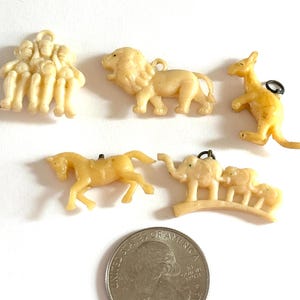 May include: A collection of small, cream-colored plastic animal charms, including a lion, kangaroo, horse, and elephants. A quarter is included for size reference. Each charm has a small loop for attaching to a necklace or bracelet.