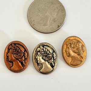 May include: Three oval-shaped cameo buttons in copper, brass, and bronze tones, featuring a profile of a woman with a laurel wreath. A US quarter coin is included for size comparison. The buttons are on a white surface.