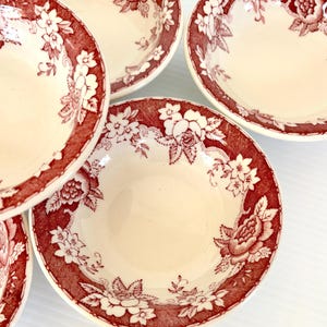 May include: A set of four round ceramic bowls featuring a cream-colored interior and a wide rim decorated with a deep red floral pattern. The design includes detailed illustrations of roses and smaller blossoms set against a textured background.