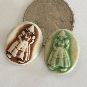 May include: Two oval ceramic pendants, one brown and one green, each featuring a Dutch girl design. A US quarter is next to the pendants. The pendants are likely for jewelry or craft projects.
