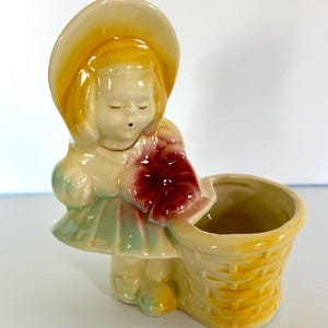 May include: A vintage ceramic figurine of a girl wearing a hat and dress, holding a red flower and a yellow basket. The figurine is painted in pastel colors, with a glossy finish. The basket is textured to resemble woven wicker.