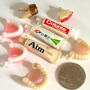 May include: A collection of miniature dental items, including teeth, dentures, and tubes of toothpaste. The toothpaste brands are Colgate, Crest, and Aim. A U.S. quarter is included for size comparison. The items are white, pink, and beige.