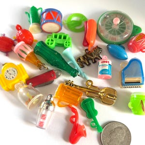 May include: A collection of miniature plastic charms and trinkets including a green bottle, a yellow clock, a red drum, a gold-tone guitar, a soup can, and a roulette wheel, arranged near a United States quarter coin for scale reference.